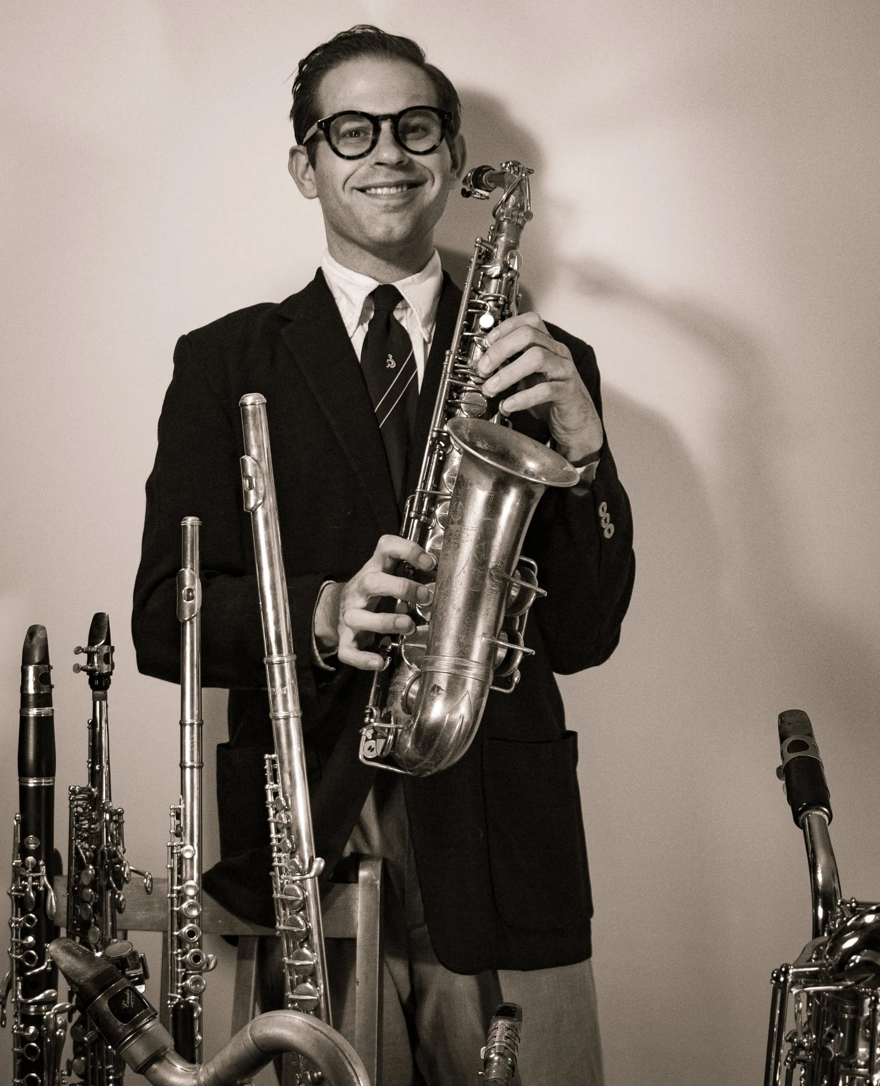 Aaron Johnson in a dark jacket and tie, smiling and holding an alto saxophone, a rack of flutes and clarinets standing around him.