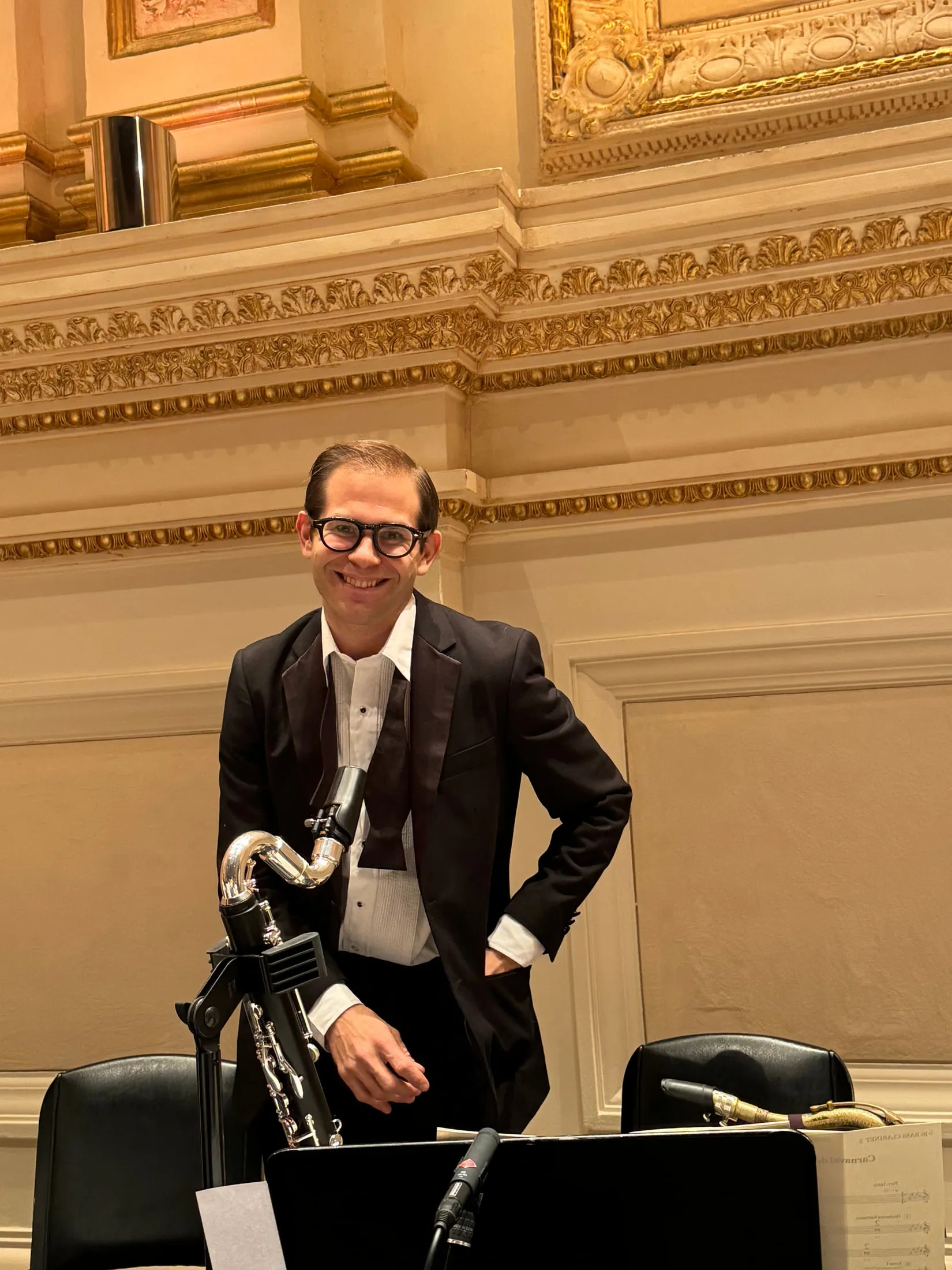 Aaron Johnson in white tie standing at his chair in a gilded concert hall, clarinets on stands in front of him.