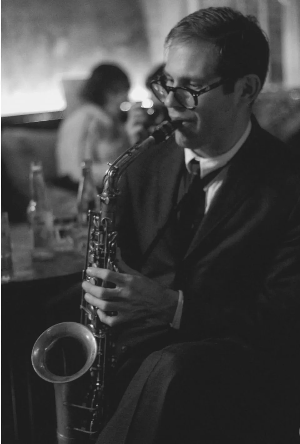 Aaron Johnson playing tenor saxophone beside a club table, photographed in black and white.