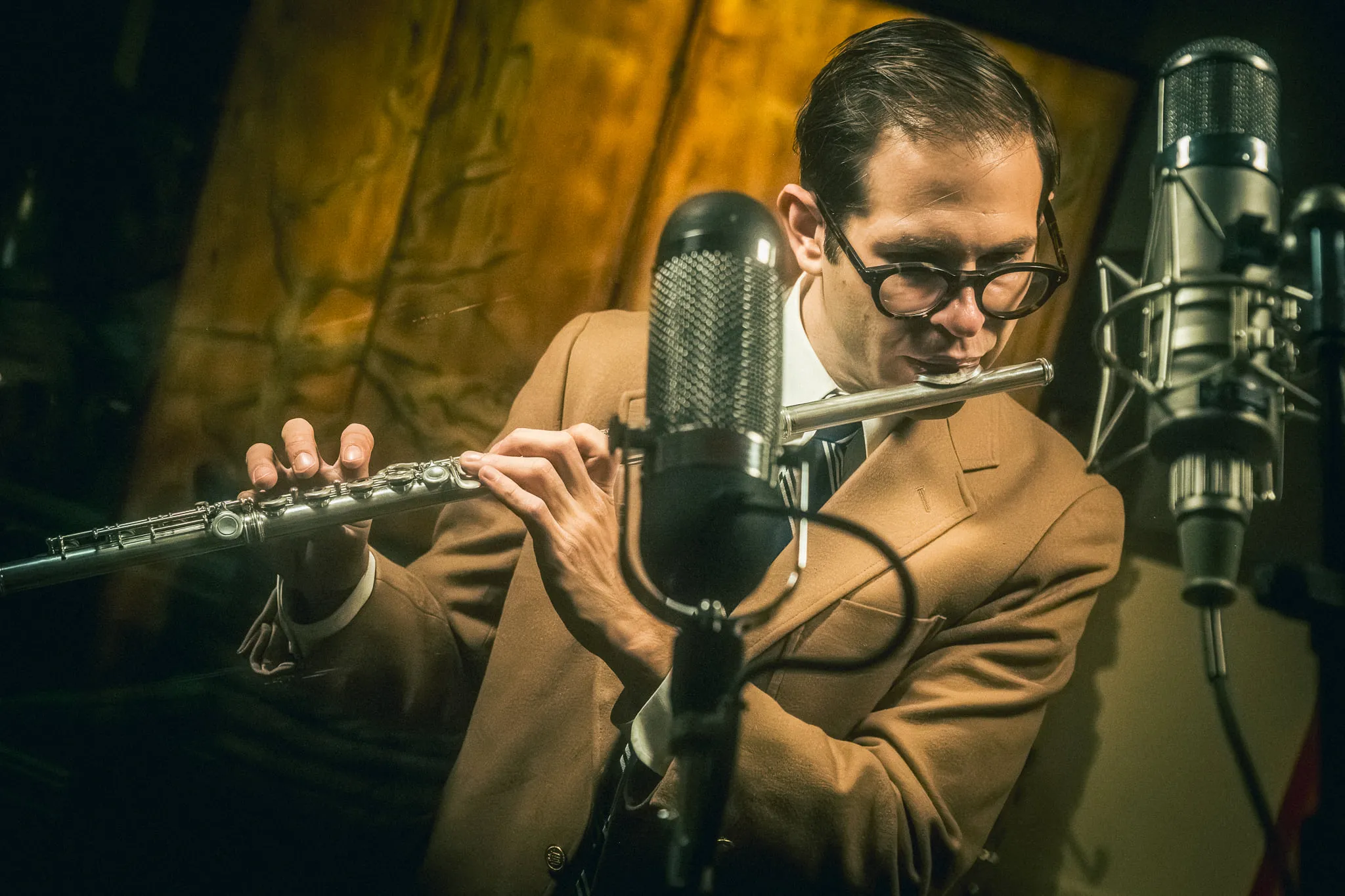 Aaron Johnson in a tan jacket playing flute between two studio microphones.