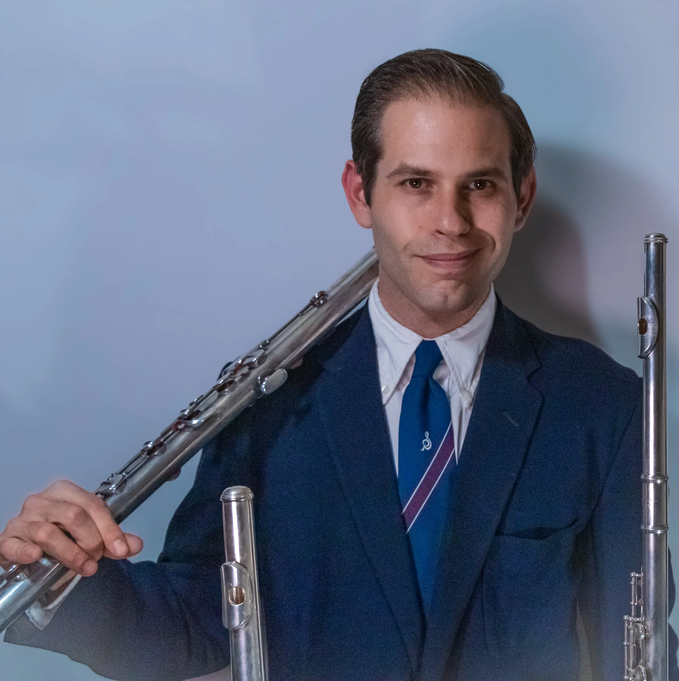 Aaron Johnson in a navy blazer and striped tie holding two flutes, against a blue-grey studio backdrop.