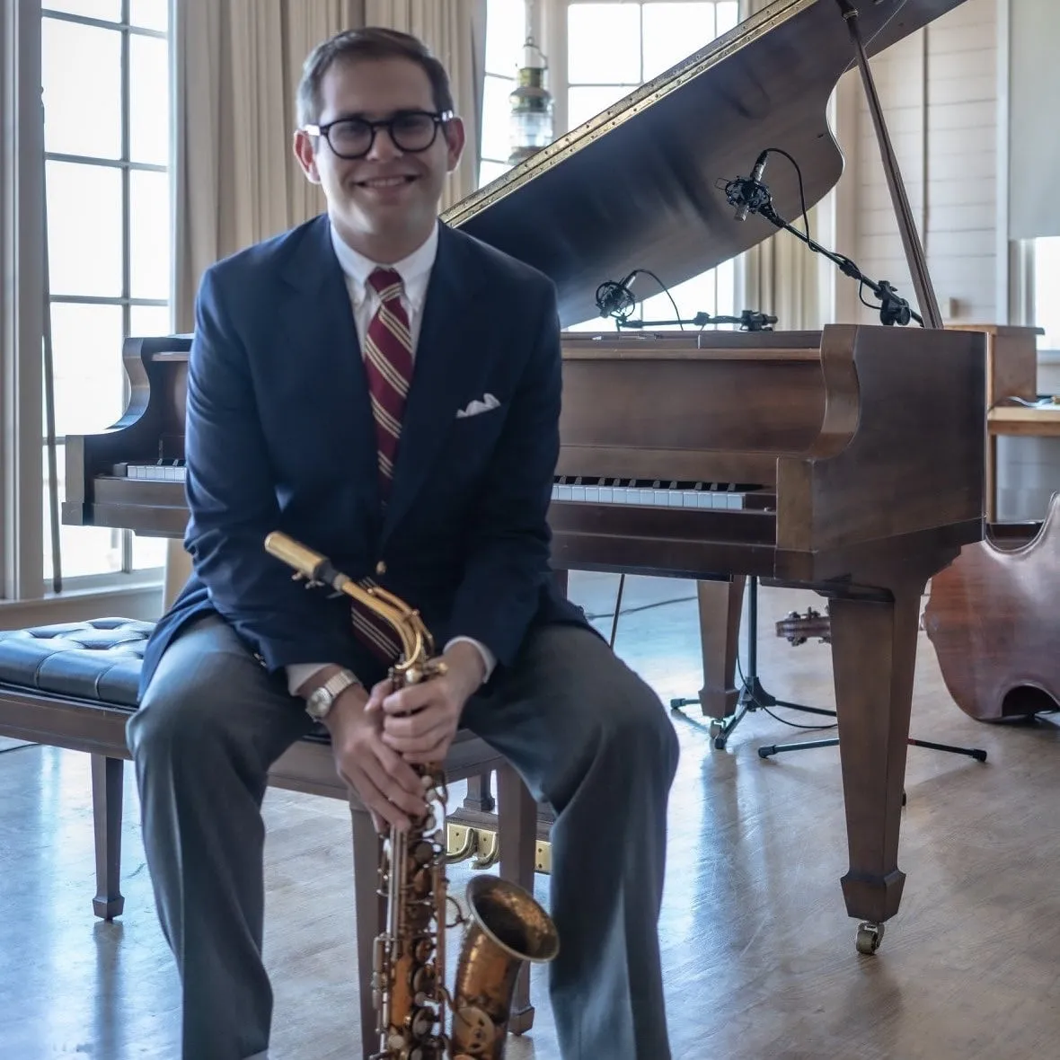 Aaron Johnson seated beside an open grand piano with an alto saxophone resting on his knee.