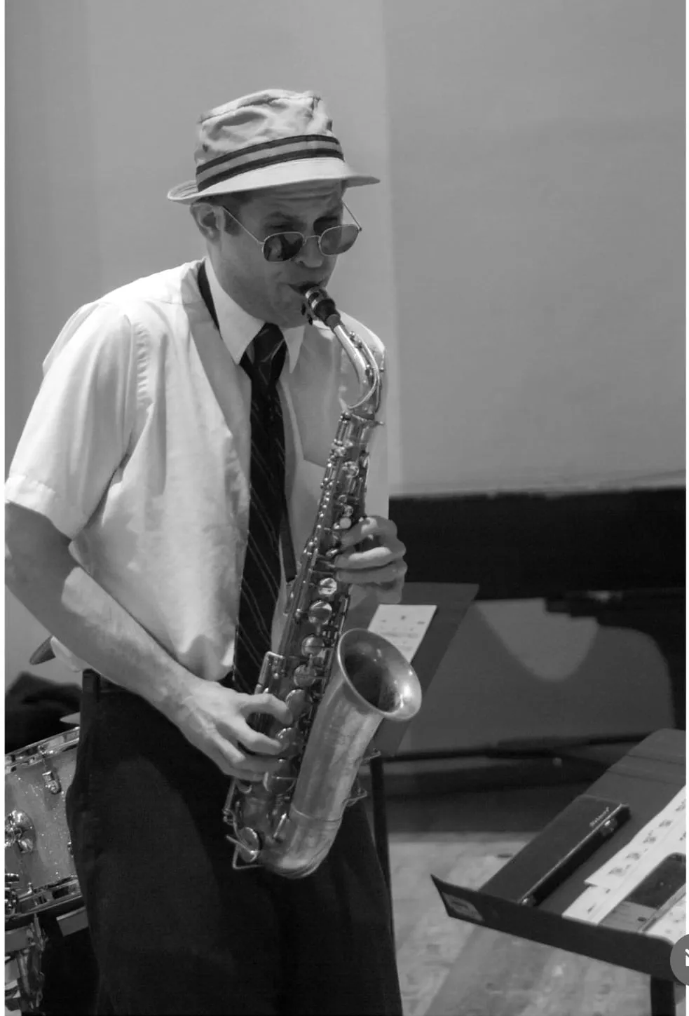 Aaron Johnson in shirtsleeves and a straw hat playing alto saxophone at a rehearsal, in black and white.