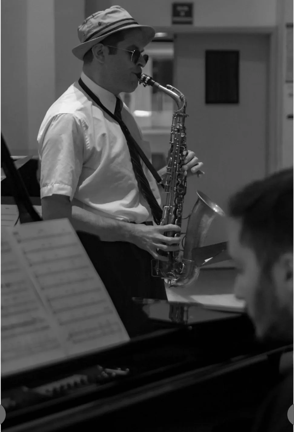 Aaron Johnson in a straw hat playing alto saxophone beside an open grand piano, in black and white.