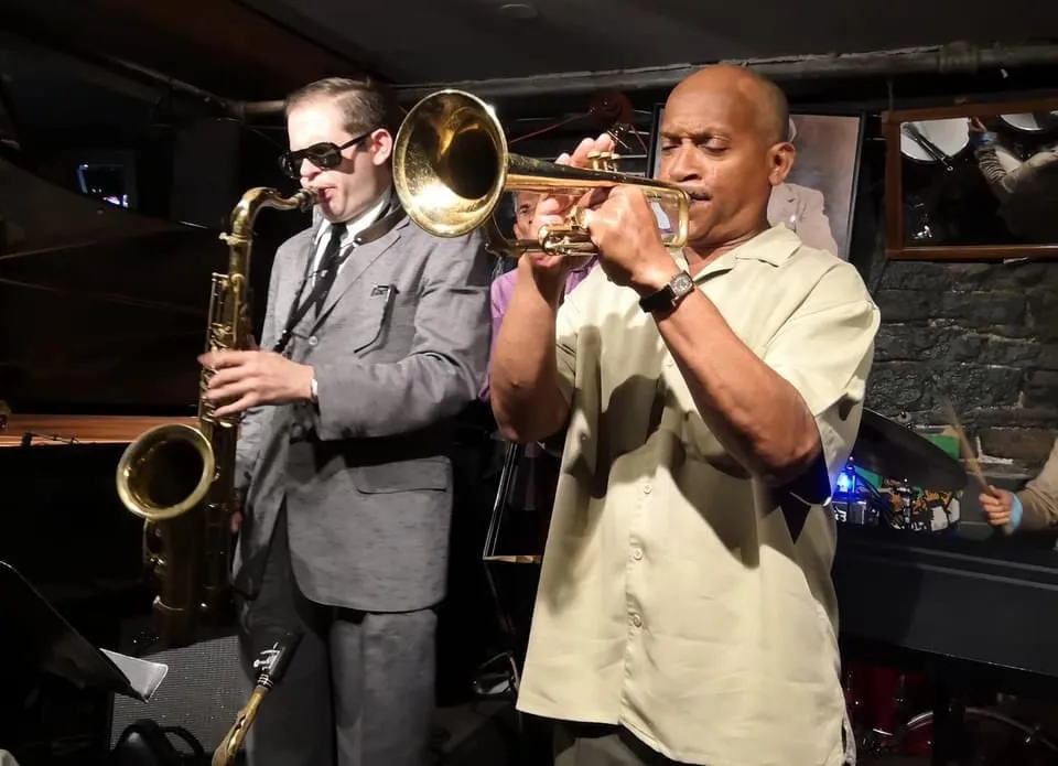 Aaron Johnson on saxophone playing a line beside a trumpeter on a club bandstand.