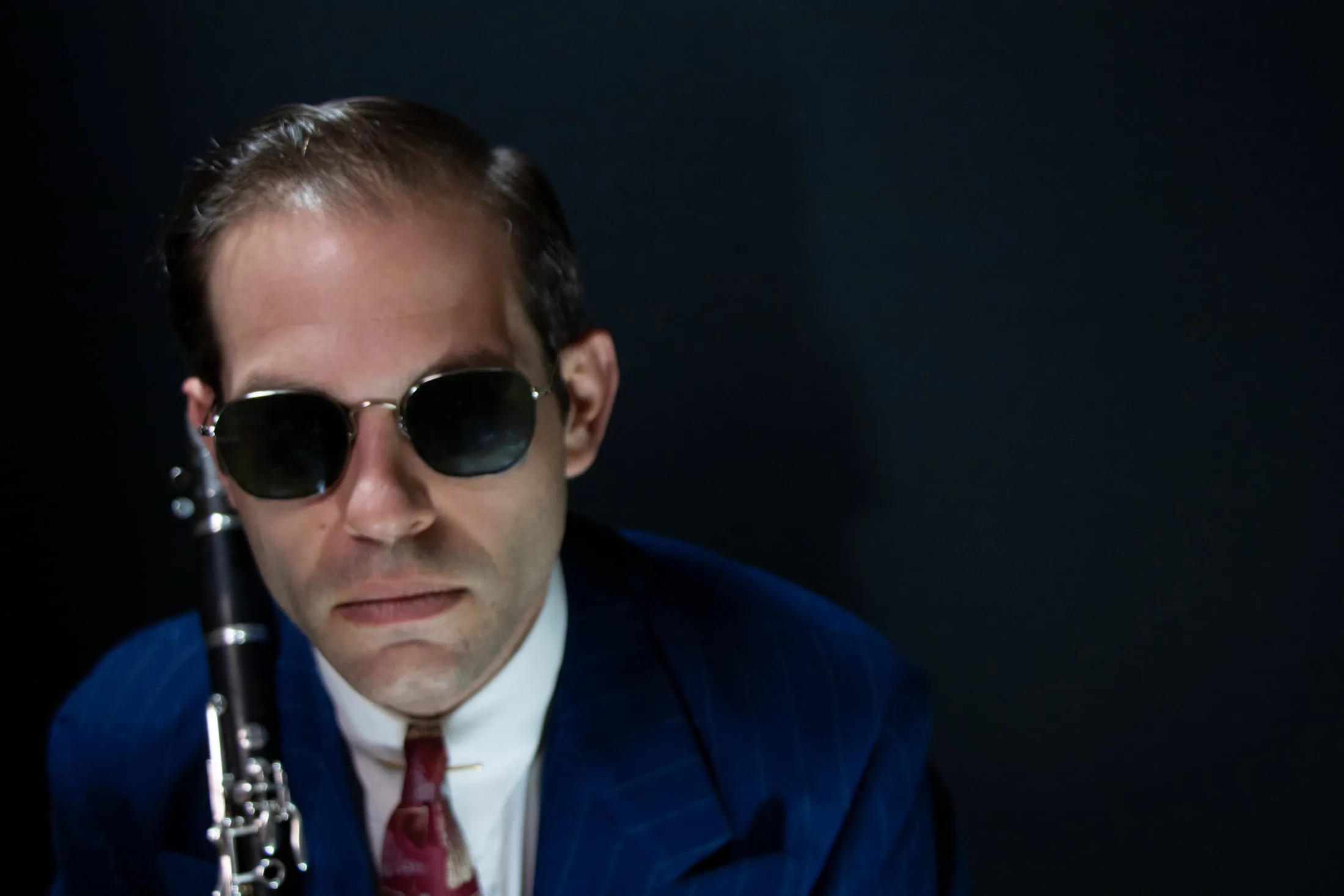 Aaron Johnson in dark sunglasses and a blue suit against a near-black background, a clarinet resting against his shoulder.