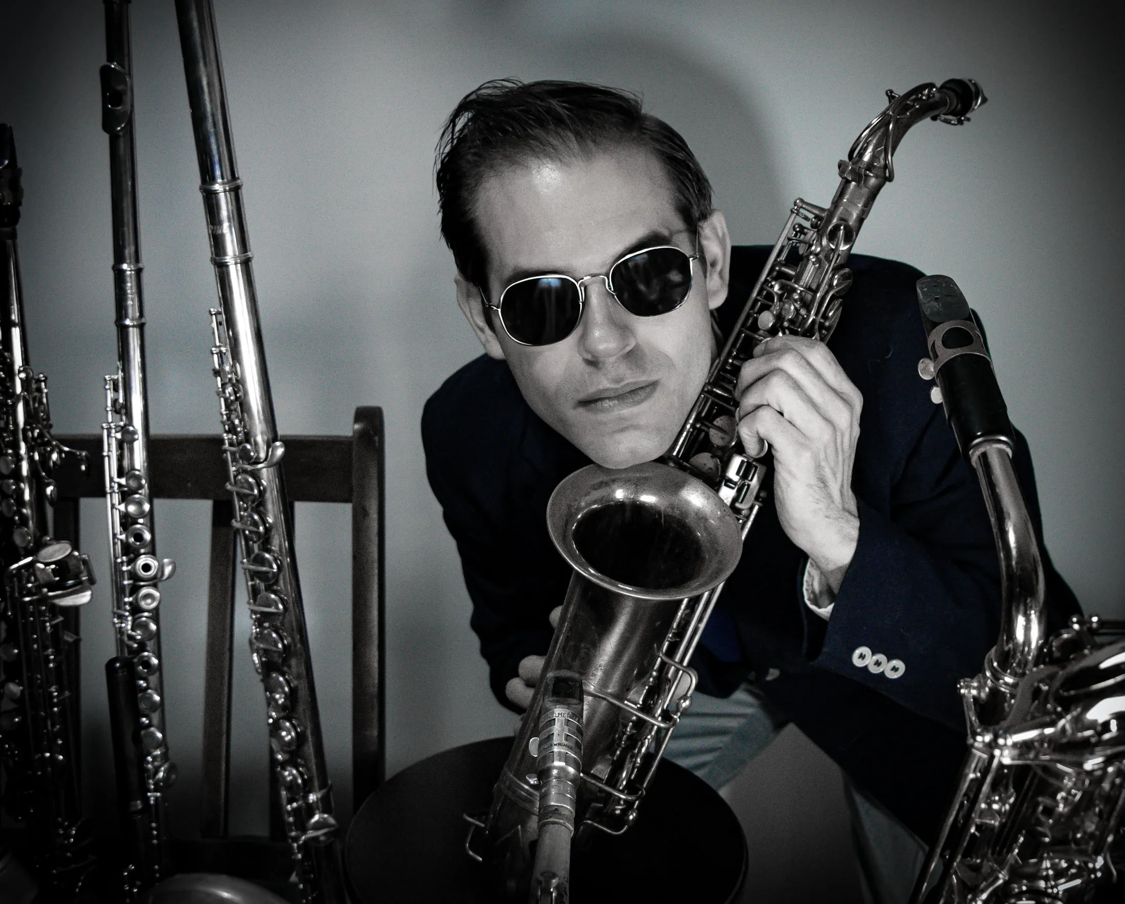 Aaron Johnson in dark glasses leaning on a tenor saxophone, a rack of woodwinds behind him.