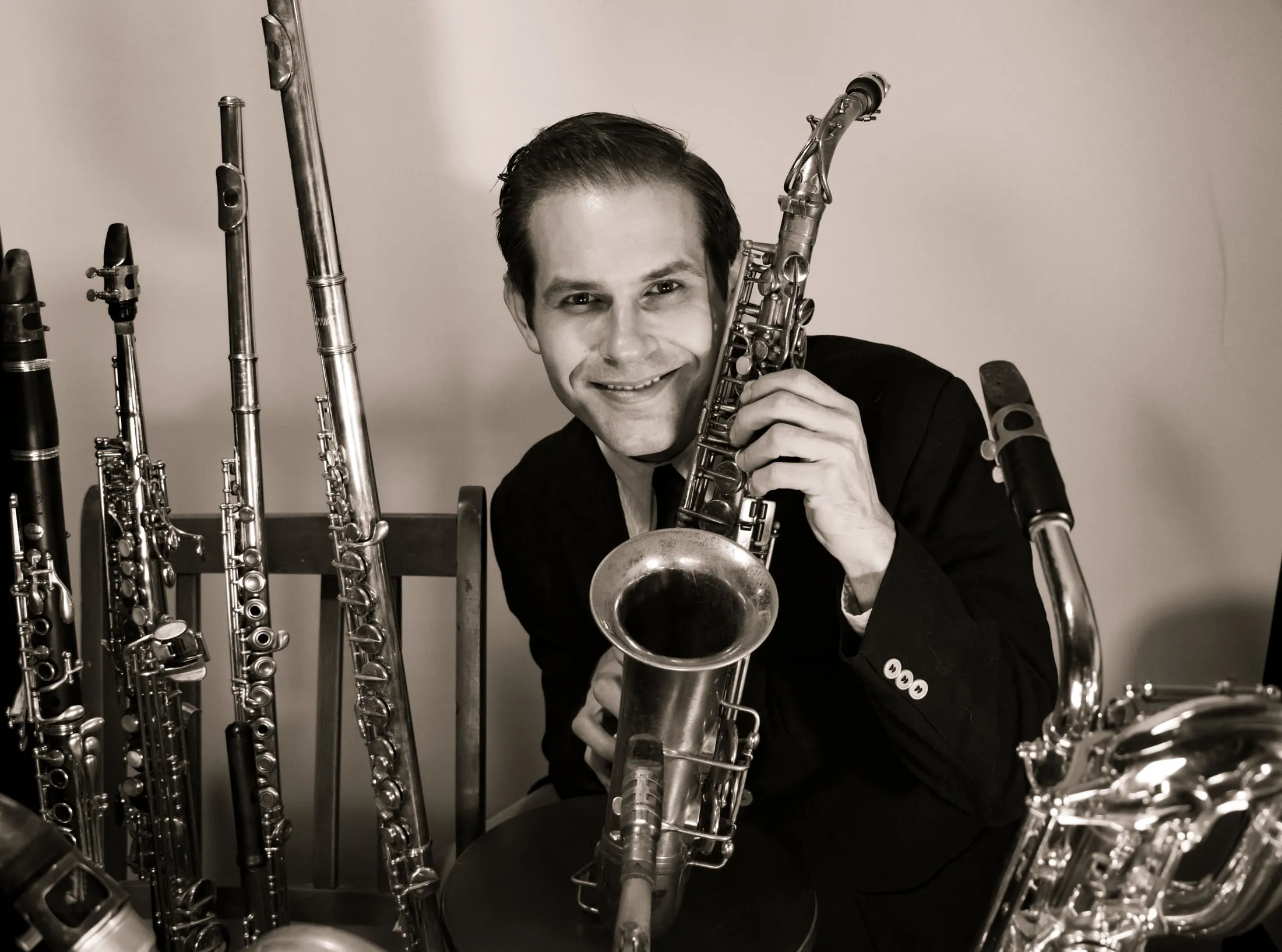 Aaron Johnson seated behind the bell of a tenor saxophone, flutes and clarinets standing behind him.