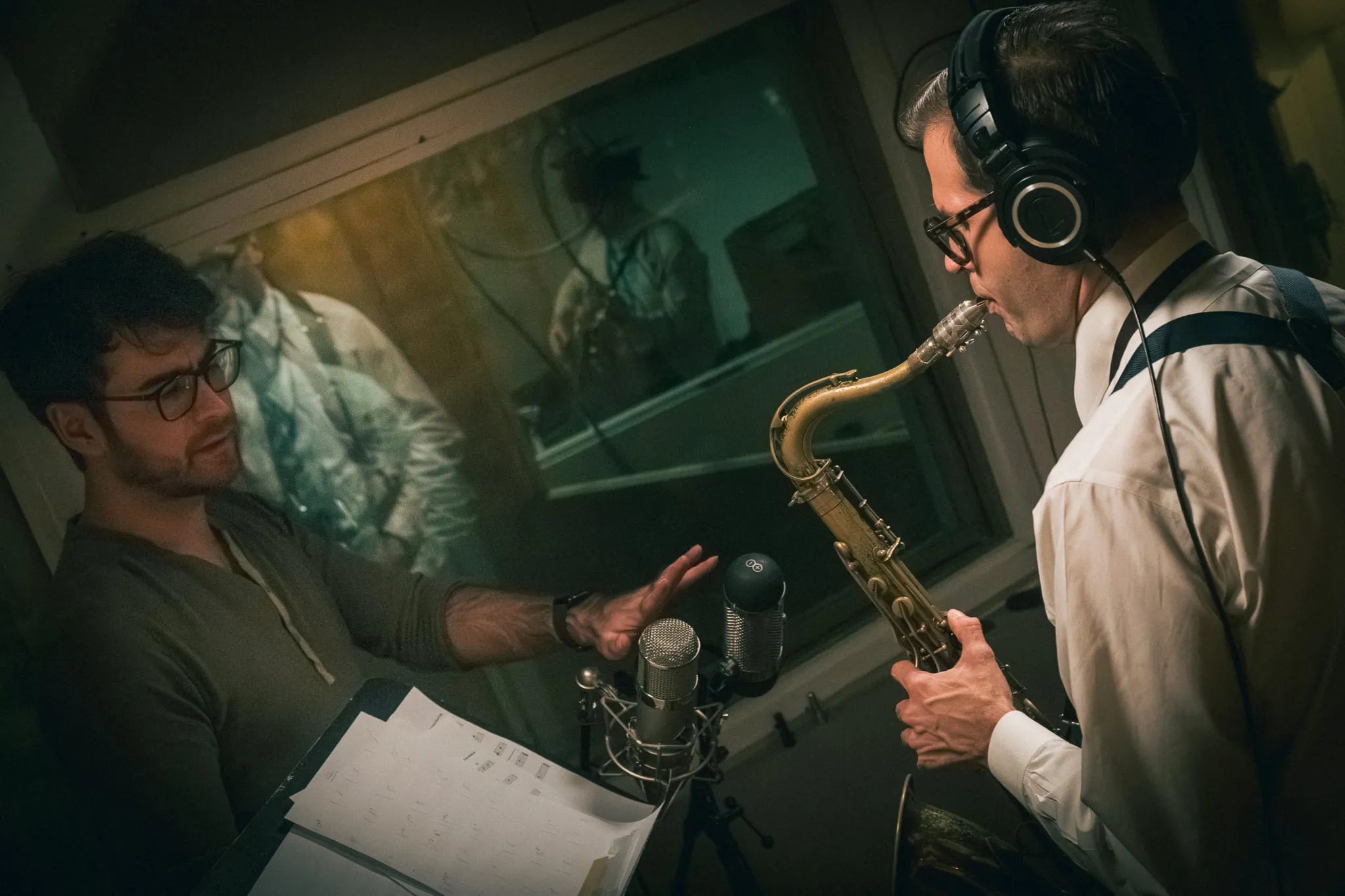 Aaron Johnson on tenor saxophone talking through the control-room glass with an engineer holding a score.